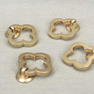 Może przedstawiać: Cztery złote metalowe gałki do szafek w kształcie koniczyny. Każda gałka ma okrągłą podstawę i wypukły kształt koniczyny z otworem w środku.