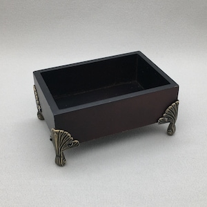May include: A small, rectangular wooden box with a dark brown finish. The box has decorative metal feet with a leaf design. The box is empty.
