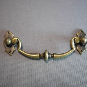 May include: A gold-toned metal drawer pull with a decorative design. The pull has a curved shape and two ornate ends.