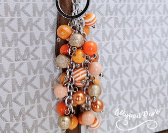 May include: A silver key chain with a large orange, peach, and gold beaded tassel. The tassel is attached to a silver ring and a silver lobster clasp. The keychain is hanging from a white and brown patterned handbag with the text "MICHAEL KORS" printed on it.