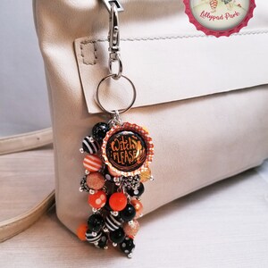 May include: A black and orange Halloween-themed keychain with a charm that says "Witch Please". The keychain is attached to a silver key ring and has a variety of beads, including black and white striped beads, orange beads, and black beads with white polka dots.
