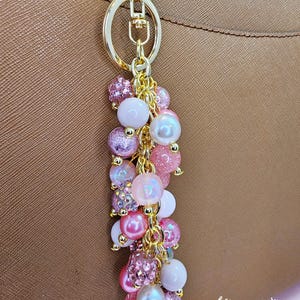 May include: A gold-toned keychain with a cluster of pink and white beads. The beads vary in size and texture, including pearls and glittery accents. The keychain is attached to a brown leather handbag.
