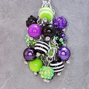 May include: A cluster of colorful beads in various shapes and sizes, including striped, solid, and crystal-covered beads. The color palette includes green, purple, black, and white, all connected by silver chains and a keyring. The text "lillypad Park" is visible.