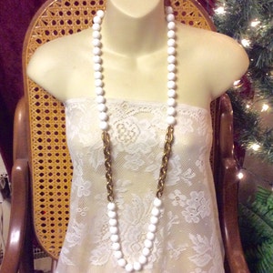 May include: A long white beaded necklace with a gold chain interwoven. The necklace is displayed on a white lace top.