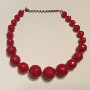 May include: A red beaded necklace with faceted beads. The necklace is made of large, round beads that are strung together on a silver chain.