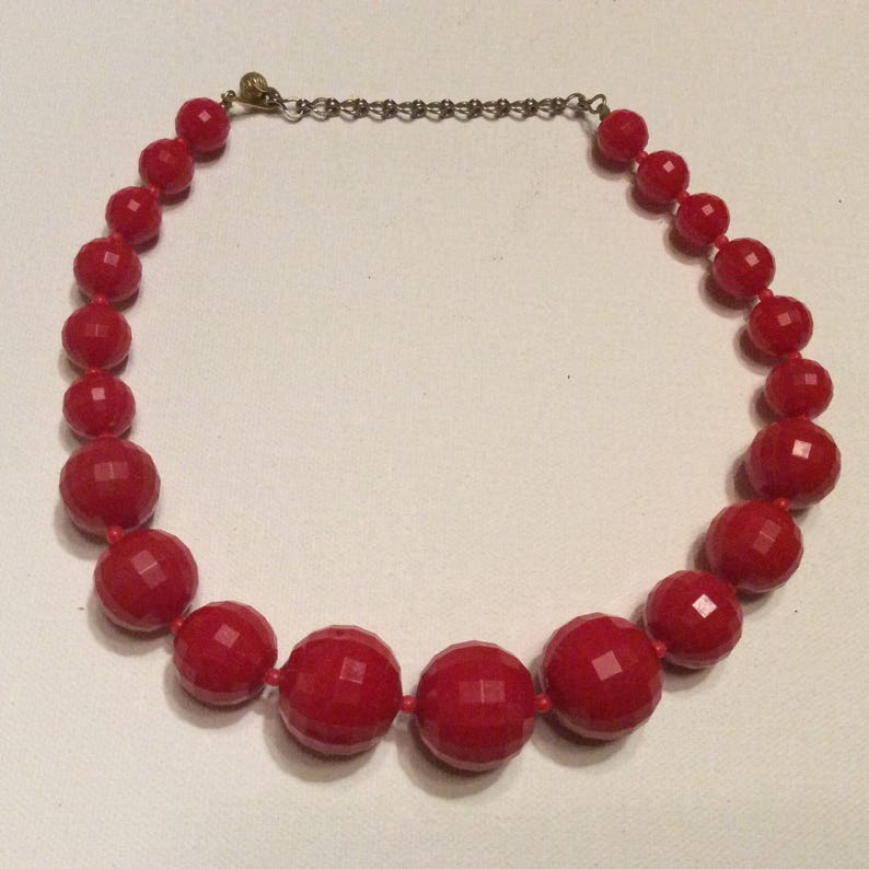 May include: A red beaded necklace with faceted beads. The necklace is made of large, round beads that are strung together on a silver chain.