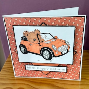 Op de afbeelding: Wenskaart met een cartoon illustratie van een teddybeer die in een oranje cabrio rijdt. De kaart heeft een patroonrand en de tekst "MINI Happy Returns" in een witte rechthoek.