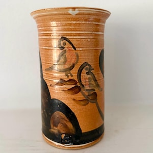 May include: A cylindrical ceramic vase with a light orange base colour, featuring horizontal ridges and hand-painted dark brown and black motifs of birds perched on branches. The vessel has a flared rim and a smooth, glossy finish throughout.