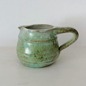 May include: A handmade ceramic jug with a rounded body and a curved handle. The jug is glazed in shades of green and brown, with visible texture and a slightly irregular shape. The spout is gently curved.
