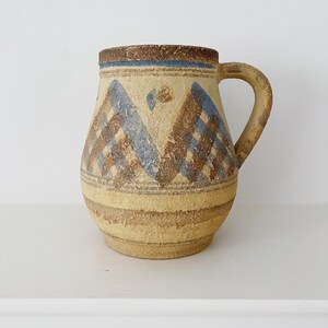May include: A beige ceramic mug with a handle, featuring blue and brown geometric patterns. The mug has a textured surface and a brown rim. The design includes horizontal stripes and triangle shapes.