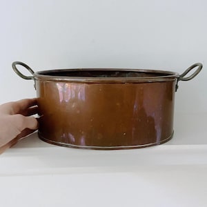 May include: A copper-coloured, oval-shaped pot with two curved handles. The pot has a shiny, reflective surface and sits on a white surface. The pot is likely used for cooking or decorative purposes.