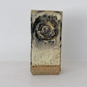 May include: A rectangular ceramic vase with a speckled cream and beige base. The upper portion features a dark, mottled glaze with a prominent circular, layered relief design in the centre, creating a textured contrast against the smooth body.