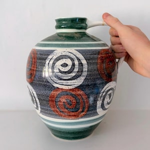 May include: A ceramic pitcher with a rounded body and a single handle. The surface features a dark blue and green mottled background with horizontal stripes and large, hand-painted spiral motifs in white and burnt orange across the exterior.