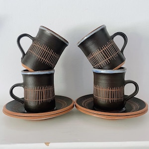 May include: Four dark brown ceramic mugs and matching saucers arranged in two stacks. Each mug features a vertical line pattern, a curved handle, and a light blue rim. The saucers have a circular etched design and a natural terracotta-coloured base.