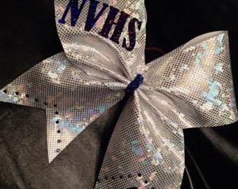 May include: A white cheer bow with a silver iridescent finish and black dots around the edges. The bow has the letters "NVHS" in dark blue on the top part of the bow.