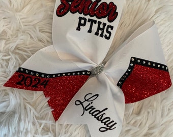 Op de afbeelding: Witte cheer bow met rode glitter en zwarte bies. De strik heeft de tekst "Senior PTHS 2024" en "Lindsay" erop.