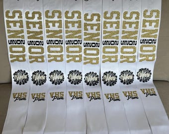 May include: Nine white satin sashes with black trim and gold glitter lettering. Each sash says "SENIOR" and has a name, "Trinity", "Macy", "Kaitlynn", "Addy", "Simane", "Sofia", "Kayleigh", and "Allyssa". Each sash also has a black and white pom-pom graphic and the text "VHS Pom".