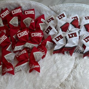 Puede incluir: Una colección de lazos de animadora rojos y blancos con detalles de purpurina plateada. Los lazos están personalizados con nombres y las iniciales de la escuela "CGHS" y "LTE".