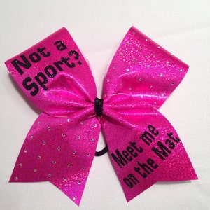 May include: Pink cheer bow with black glitter and rhinestones. The bow has the text "Not a sport?" on one side and "Meet me on the Mat" on the other side.
