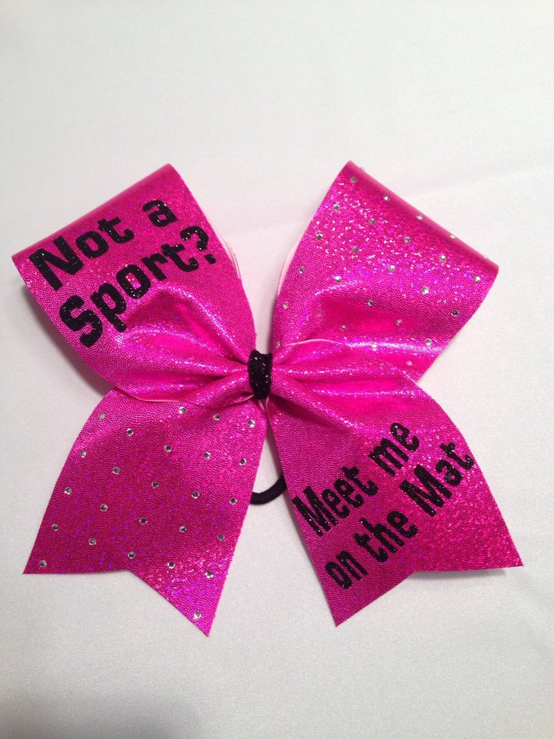 May include: Pink cheer bow with black glitter and rhinestones. The bow has the text "Not a sport?" on one side and "Meet me on the Mat" on the other side.