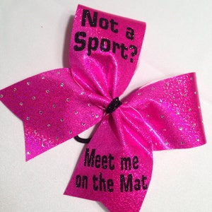 May include: Pink cheer bow with black glitter and rhinestones. The bow has the text "Not a Sport? Meet me on the Mat" printed on it.