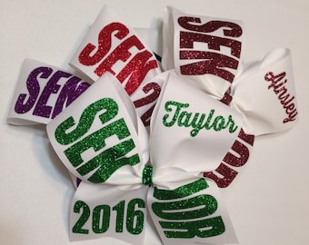 May include: White cheer bow with green glitter lettering that says "Senior 2016" and "Taylor". There are also three other bows with red, purple, and green glitter lettering that say "Senior".