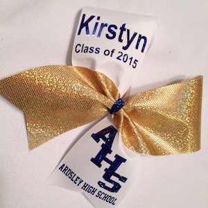 May include: A white cheer bow with gold glitter ribbon and blue and white lettering. The bow has the text "Kirstyn Class of 2015" and "Ardsley High School" with the school initials "AHS" in blue.