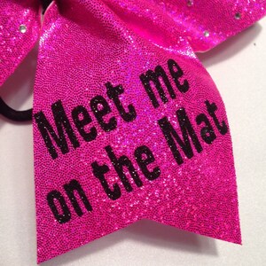 May include: A pink sparkly cheer bow with black text that reads "Meet me on the Mat".