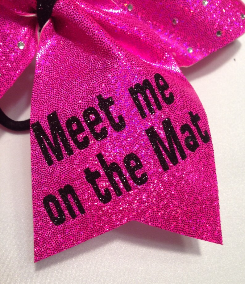 May include: A pink sparkly cheer bow with black text that reads "Meet me on the Mat".