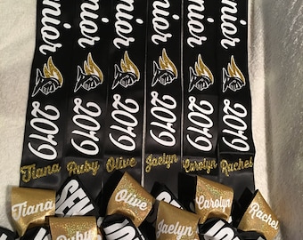 May include: Six black graduation sashes with gold glitter lettering. The sashes say "FHS Senior 2019" and each sash has a different name: Tiana, Ruby, Olive, Jaclyn, Carolyn, and Rachel.  There are also six gold and black graduation bows with the same names.