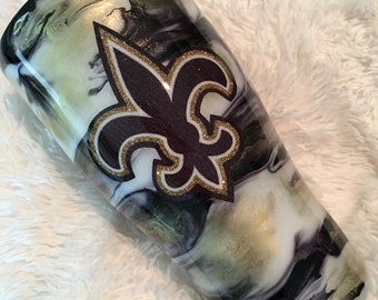 May include: A white, black and gold marbled tumbler with a gold and purple fleur-de-lis design.