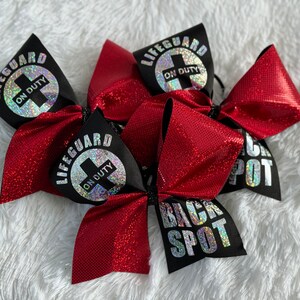 Könnte beinhalten: Drei rote und schwarze Cheer-Schleifen mit silbernem Glitzer. Eine Schleife hat den Text "Lifeguard On Duty" mit einem silbernen Kreuz. Eine andere Schleife hat den Text "Back Spot" und die dritte Schleife hat den Text "Lifeguard On Duty" mit einem silbernen Kreuz.