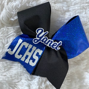 May include: A blue and black cheer bow with white glitter lettering. The bow has the text "JCHS" and "Yanet" on it.