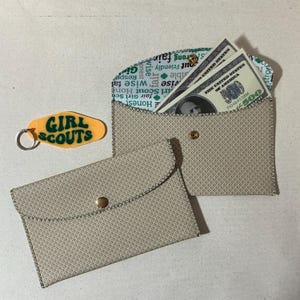 May include: Two beige fabric envelopes with a diamond pattern. The envelope on the right has a flap that is folded over and secured with a gold button. The flap is lined with a white fabric with green text that reads "Honest, fair Girl Scout Home, wise fair, wise, out Friendly, strong fair, monsibla anato, Resp, Gi". The envelope on the left has a flap that is folded over and secured with a gold button. The envelope is empty.
