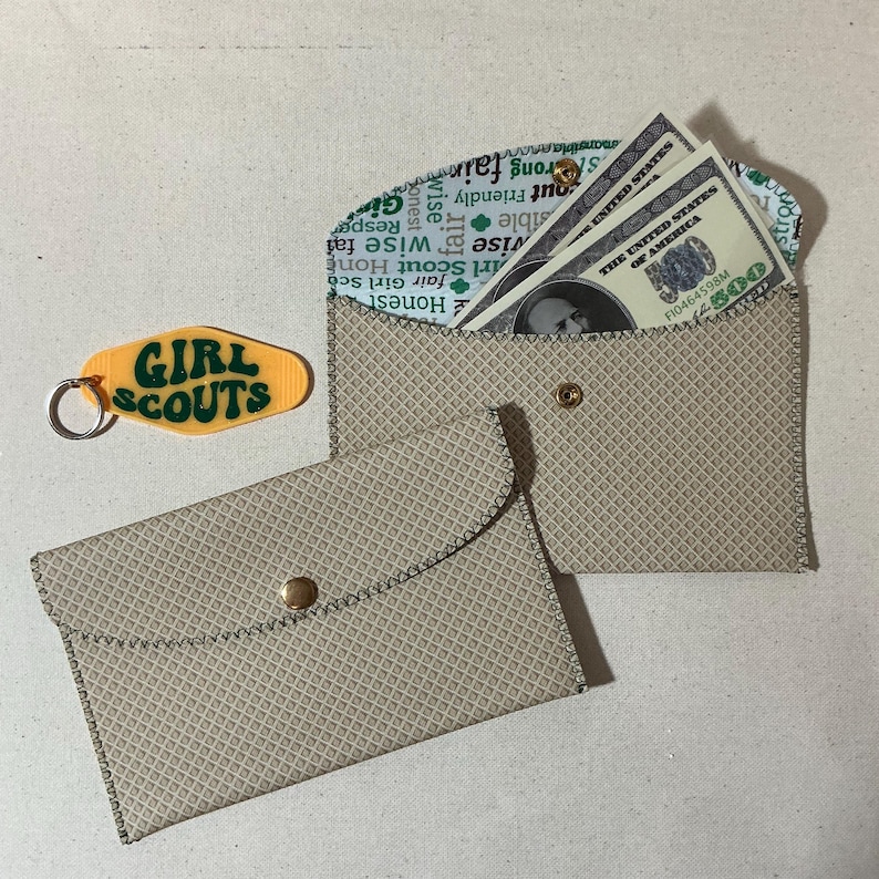 May include: Two beige fabric envelopes with a diamond pattern. The envelope on the right has a flap that is folded over and secured with a gold button. The flap is lined with a white fabric with green text that reads "Honest, fair Girl Scout Home, wise fair, wise, out Friendly, strong fair, monsibla anato, Resp, Gi". The envelope on the left has a flap that is folded over and secured with a gold button. The envelope is empty.