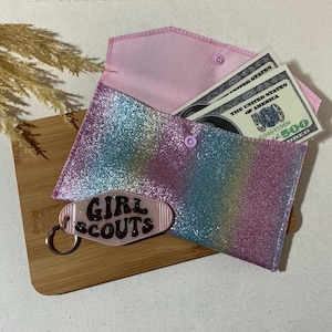 May include: A rainbow glitter envelope with a pink felt lining and a snap closure. The envelope has a key chain attached with a pink plastic tag that says "Girl Scouts".