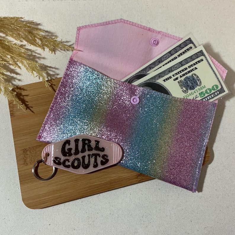 May include: A rainbow glitter envelope with a pink felt lining and a snap closure. The envelope has a key chain attached with a pink plastic tag that says "Girl Scouts".