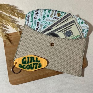 May include: A beige fabric wallet with a zippered closure and a Girl Scouts keychain. The wallet has a green and white pattern with the words "Girl Scouts" repeated. The keychain is yellow with green lettering and a sparkly finish.