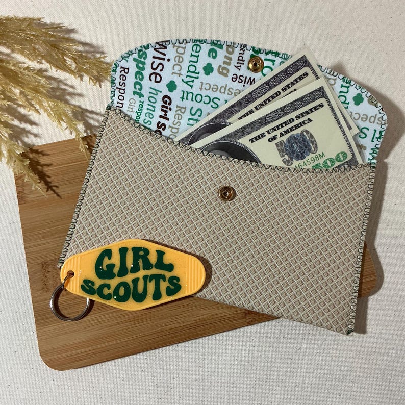 May include: A beige fabric wallet with a zippered closure and a Girl Scouts keychain. The wallet has a green and white pattern with the words "Girl Scouts" repeated. The keychain is yellow with green lettering and a sparkly finish.