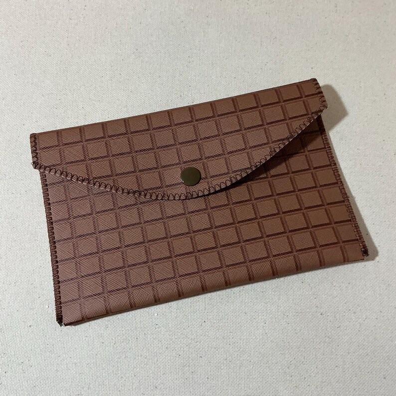 May include: A brown envelope with a flap closure and a single button. The envelope has a checkered pattern.