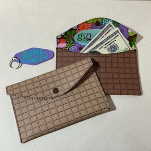 May include: Two brown fabric wallets with a grid pattern. The top wallet is open and has a floral fabric lining with a Girl Scout patch and US dollar bills inside. The bottom wallet has a snap closure.