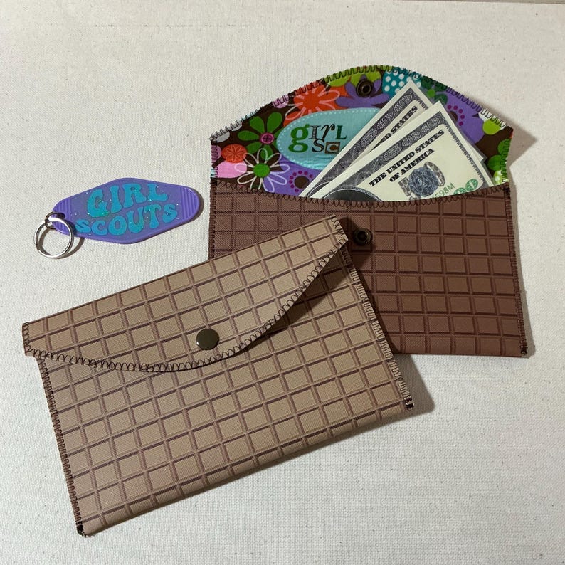 May include: Two brown fabric wallets with a grid pattern. The top wallet is open and has a floral fabric lining with a Girl Scout patch and US dollar bills inside. The bottom wallet has a snap closure.