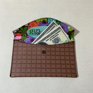 May include: A brown fabric envelope with a checkered pattern and a snap closure. The envelope is open and has several dollar bills inside. The inside of the envelope is lined with a colorful floral fabric with the word "Girl" printed on it.