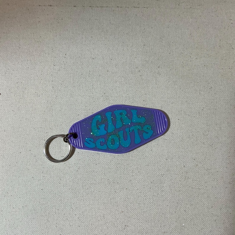 May include: Purple plastic keychain with a silver ring. The keychain is shaped like a motel key and has the words "GIRL SCOUTS" in teal glitter letters.