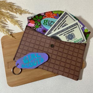 May include: A brown wallet with a checkered pattern and a zipper closure. The wallet has a purple keychain with the words "Girl Scouts" on it. The wallet is open and has several US dollar bills inside.