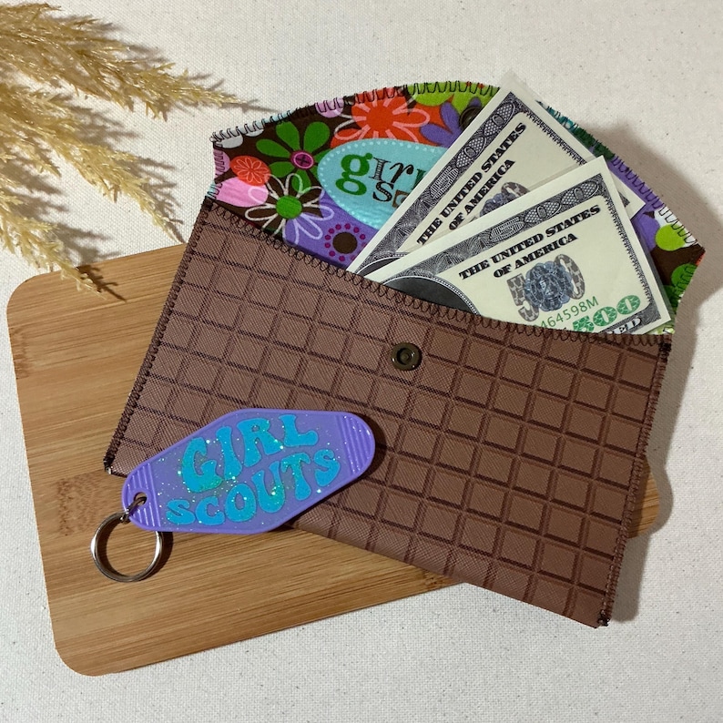 May include: A brown wallet with a checkered pattern and a zipper closure. The wallet has a purple keychain with the words "Girl Scouts" on it. The wallet is open and has several US dollar bills inside.