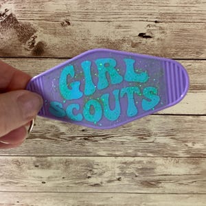 May include: Purple motel key chain with the words "GIRL SCOUTS" in turquoise glitter lettering.