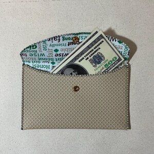 May include: A beige fabric envelope with a green and white patterned lining. The envelope has a snap closure and a dollar bill is visible inside.