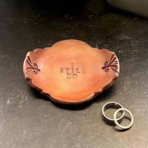 May include: A small, brown ceramic ring dish with the words "I STILL DO" stamped in the center. Two gold wedding rings sit next to the dish. The dish has a decorative edge.