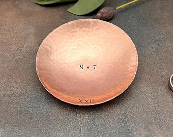 May include: A hammered copper ring dish with the initials "N + T" and the Roman numeral "XXII" engraved on it. Two rings are placed next to the dish. The background features white flowers and greenery.